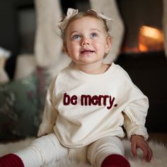 A young child with bright blue eyes and a soft smile sits on a cozy white rug, wearing a cream-colored sweater with the words "be merry" in textured red lettering. They have a bow in their hair, and a warm, festive ambiance fills the background. Gingerbread Embroidery, Girls Outerwear, Be Merry, Holiday Wardrobe, Gentle Touch, Boys Pajamas