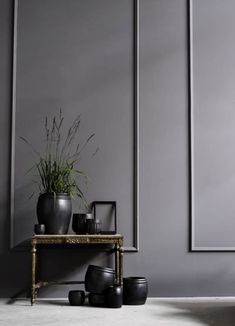 there is a table with plants on it in front of a gray wall and some black vases