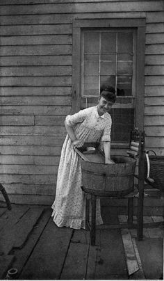 an old black and white photo of a woman washing her hands in a bucket on the porch