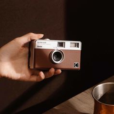 a person holding an old fashioned camera in their hand next to a metal cup on a table