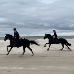 two people riding horses on the beach in front of an ocean and cloudy sky at dusk