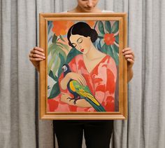 a woman holding up a painting with a bird on it's arm and the image is framed in wood