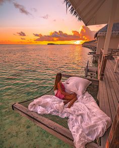 a woman laying on top of a bed next to the ocean at sunset in front of a pier