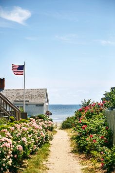 a path leading to the beach with flowers on both sides and an american flag flying in the background