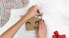 someone cutting out a paper reindeer ornament on a white table with red and green ribbon