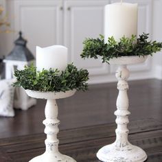 two white candles sitting on top of a table with greenery in the centerpieces