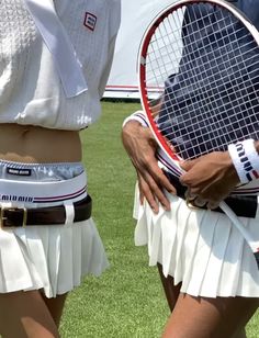 two women holding tennis racquets on top of a grass covered field with people in the background