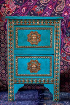 an ornate blue and gold painted chest of drawers on a carpeted area with paisley wallpaper