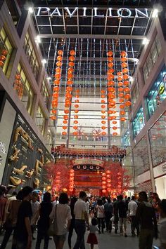 many people are walking around in front of a building with orange lanterns hanging from the ceiling