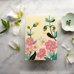 a card with flowers on it sitting next to some white and pink flowers in the background