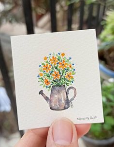 a person holding up a card with flowers in a watering can on it's front