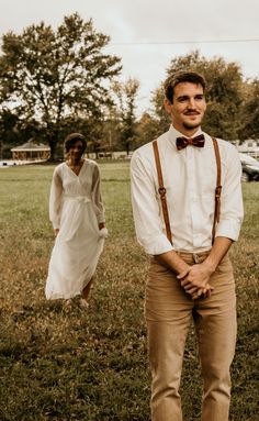 a man wearing suspenders and a bow tie standing next to a woman in a field