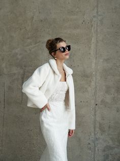 a woman in a white dress and jacket with sunglasses on her head, standing against a concrete wall