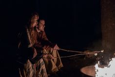 two people sitting around a campfire in the dark, one is holding a rope