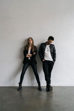 two people standing next to each other in front of a white wall with one person leaning against the wall