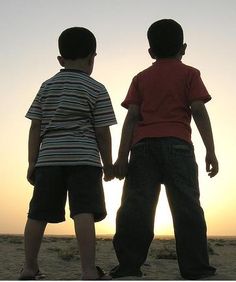 two young boys standing on top of a sandy beach next to the ocean at sunset