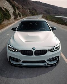 the front end of a white bmw car driving on a road near water and hills