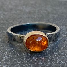 An organic, handcrafted mixed metals ring with a bright orange glowing Mandarin Spessartite Garnet. Size of the smooth polished Mandarin Spessartite Garnet cabochon is 9 x 7 mm, and it is set in a setting with side walls of 14k Gold and base of Sterling Silver. The approximately 3 mm wide ring band has been gently hammered and given an oxidized dark patina to create an organic 'snake skin' look. Ring size is US 8-8.5. Worldwide free shipping. Orange Citrine Jewelry With Polished Finish, Orange Fine Jewelry With Polished Finish, Orange Oval Jewelry With Polished Finish, Oval Orange Polished Jewelry, Orange Polished Jewelry For Anniversary, Modern Orange Gemstone Rings, Modern Orange Oval Rings, Unique Oval Orange Jewelry, Modern Orange Rings For Gifts