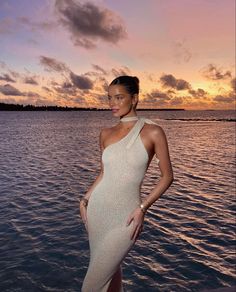 a woman standing on the edge of a body of water wearing a one shoulder dress