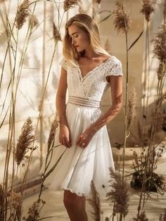 a woman in a white dress standing next to some tall grass and dried flowers with her hands on her hips