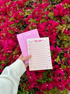 a person holding up a to do list in front of pink bougaia flowers