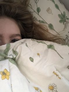 a woman laying in bed under a blanket with flowers on it and looking at the camera