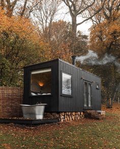 a small black house with a tub in the front yard and trees surrounding it on an autumn day