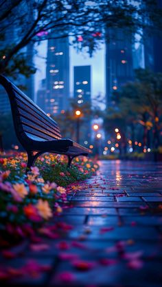 a park bench sitting in the middle of a flower garden at night with buildings in the background