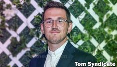 a man wearing glasses and a suit standing in front of a wall with green plants