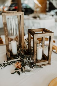 two wooden lanterns with white candles and greenery on the table at a wedding reception