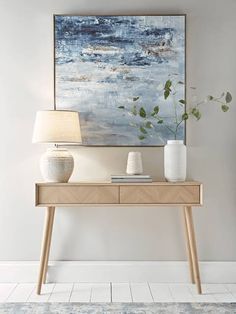 a white vase sitting on top of a wooden table next to a lamp and painting