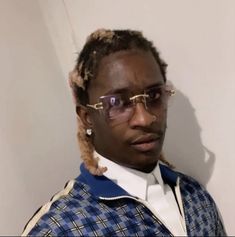 a man wearing glasses and a plaid shirt with braids on his hair is standing in front of a white wall