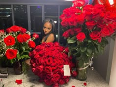 a woman is sitting in front of roses and other flowers on a counter top with a large vase full of red roses