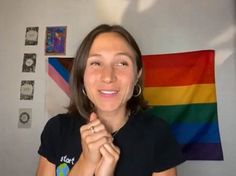 a woman standing in front of a rainbow flag and holding her hands up to her chest