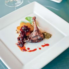 a white square plate topped with meat and veggies next to wine glasses on a table