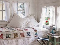 a bed with white sheets and pillows sitting next to two windows in a room filled with books