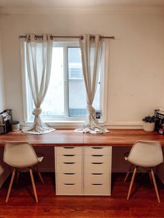 two white chairs sitting at a desk in front of a window with curtains on the windowsill