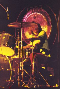 a man sitting in front of a drum set on top of a stage with lights behind him