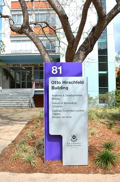 a purple sign sitting next to a tree on top of a grass covered field in front of a building