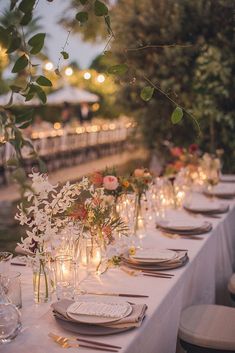 a long table is set with white linens and place settings for an outdoor dinner