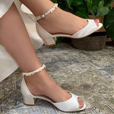 a woman wearing white high heel shoes with ankle straps and pearls on the side, sitting in front of a potted plant