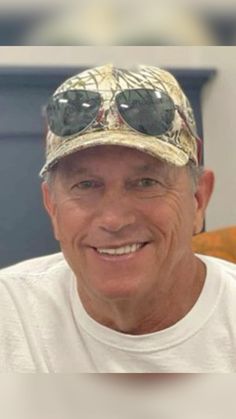 an older man wearing sunglasses and a camo hat smiles at the camera while sitting in a chair