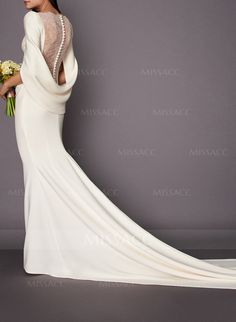 a woman in a long white dress with flowers on her shoulder and back, standing against a gray background