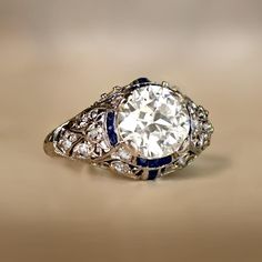 This Art Deco engagement ring centers a 2.24 carat old European cut diamond, K color and VS2 clarity. The center diamond is set in prongs and circled by a halo of calibre French cut natural sapphires. The mounting of this ring is handcrafted platinum and features floral open-work filigree, adorned with single-cut diamonds and fine milgrain. The openwork design continues down the shoulders and along the under-gallery, which is also set with sapphires. Hand-engravings decorate the shank. This ring Antique Diamond And Sapphire Engagement Ring, Vintage Looking Engagement Rings, Diamond And Sapphire Engagement Ring, Engament Rings, Sapphire Engagement Ring Halo, K Color Diamond, Estate Diamond Jewelry, Fav Products, Sapphire Rings