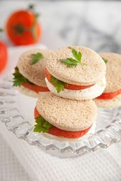 three small sandwiches on a glass plate with tomatoes in the background