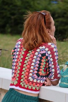 a woman with red hair wearing a colorful crochet sweater and green skirt sitting on a bench