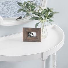 a white table with a small wooden frame on it and a potted plant in front of the mirror