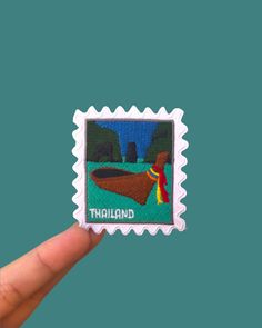 a hand holding up a stamp with the image of a boat on it and trees in the background