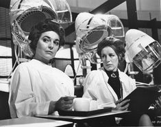 two women sitting at a table with cups and plates in front of them, one reading a book