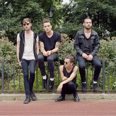 four young men are sitting on a bench in front of some bushes and trees, one is looking at the camera